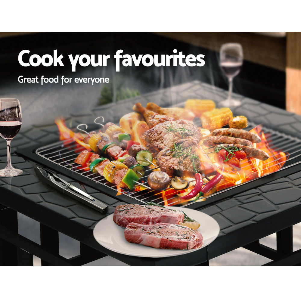 A black 2-in-1 steel fire pit/BBQ grill outdoor table with a geometric pattern on the tabletop with food being grilled, steaks on a plate ready to cook and a couple of glasses of wine on the tabletop surround.