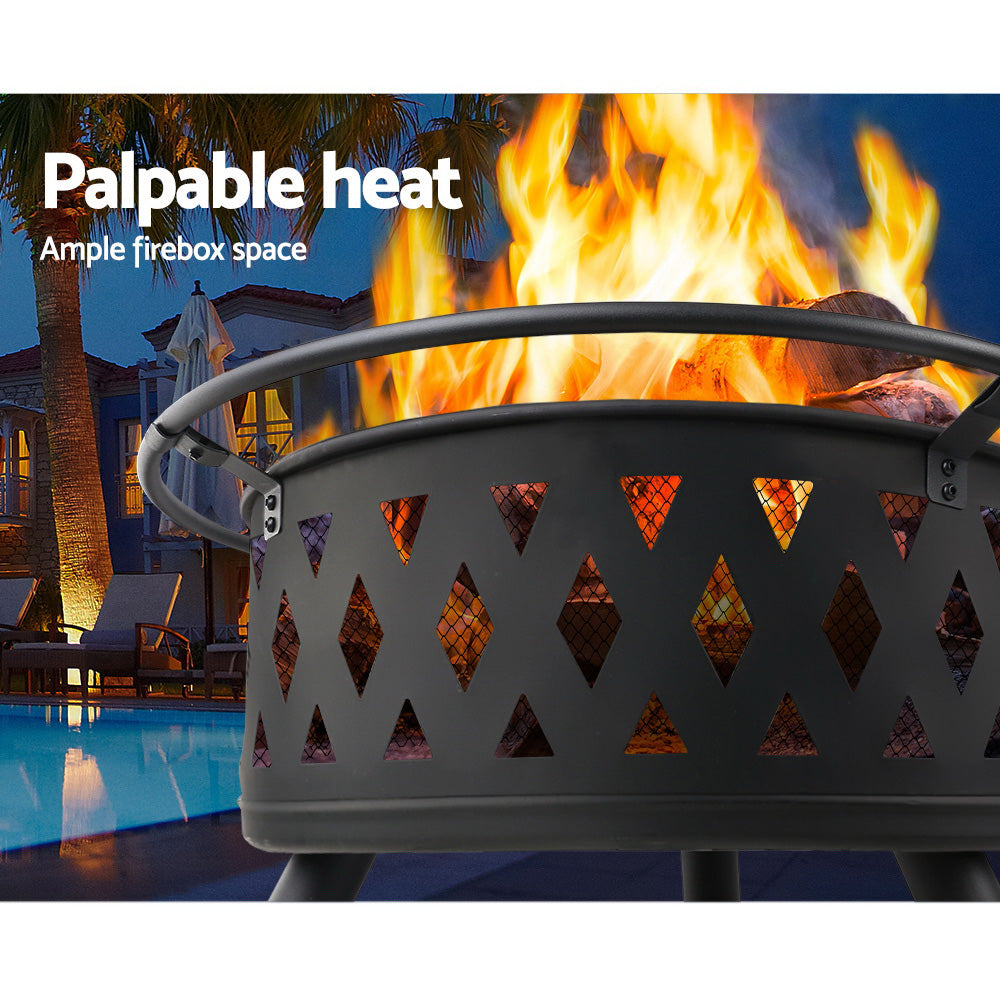 Close-up image of a black steel fire pit BBQ grill with a fire burning at a poolside in front of a house and palm trees in the nighttime.
