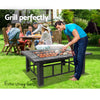 A man bending over meat that he is cooking on an outdoor fire pit BBQ grill on a black steel frame with ceramic tiles in a garden setting with family gathered around a picnic table and chairs behind and a house and garden in the background.