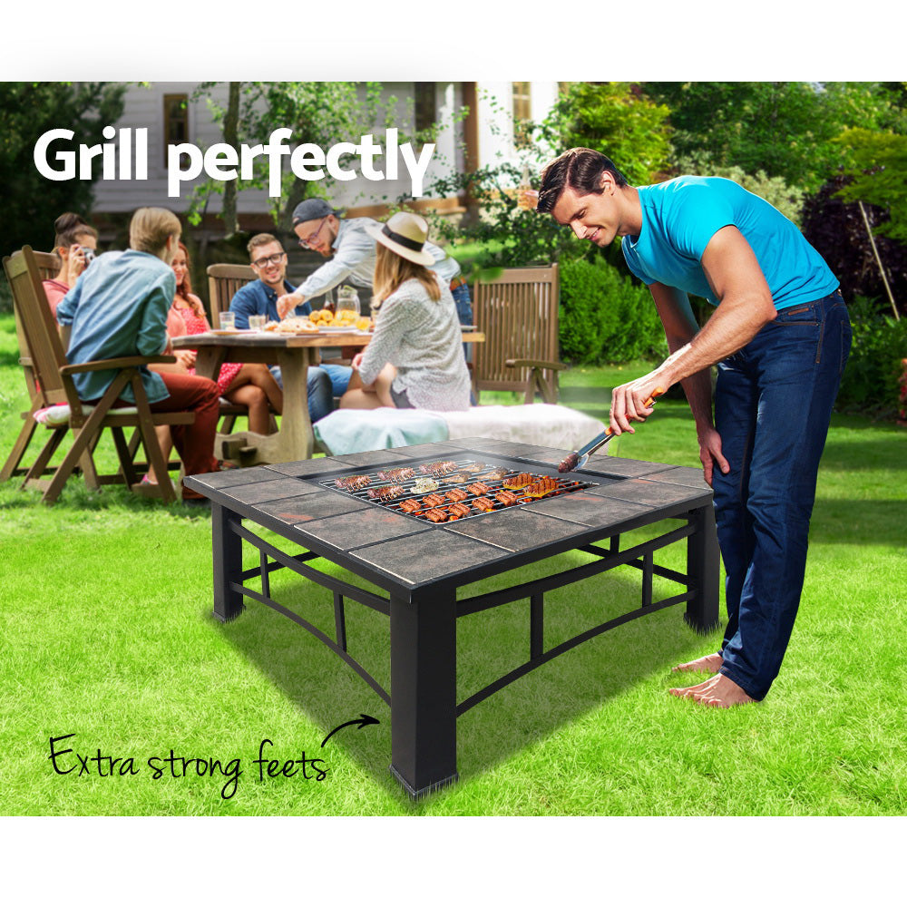 A man bending over meat that is cooking on an outdoor fire pit BBQ grill on a black steel frame with ceramic tiles in a garden setting with family gathered around a picnic table and chairs behind and a house and garden in the background.