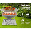A portable stainless steel Grillz 2-in-1 Fire Pit BBQ Grill, cooking steaks, sausages and vegetables with labels showing that it is stainless steel, portable, 8.5kg weight and adjustable height. Set on green grass in a garden setting with plants and slate wall in the background.
