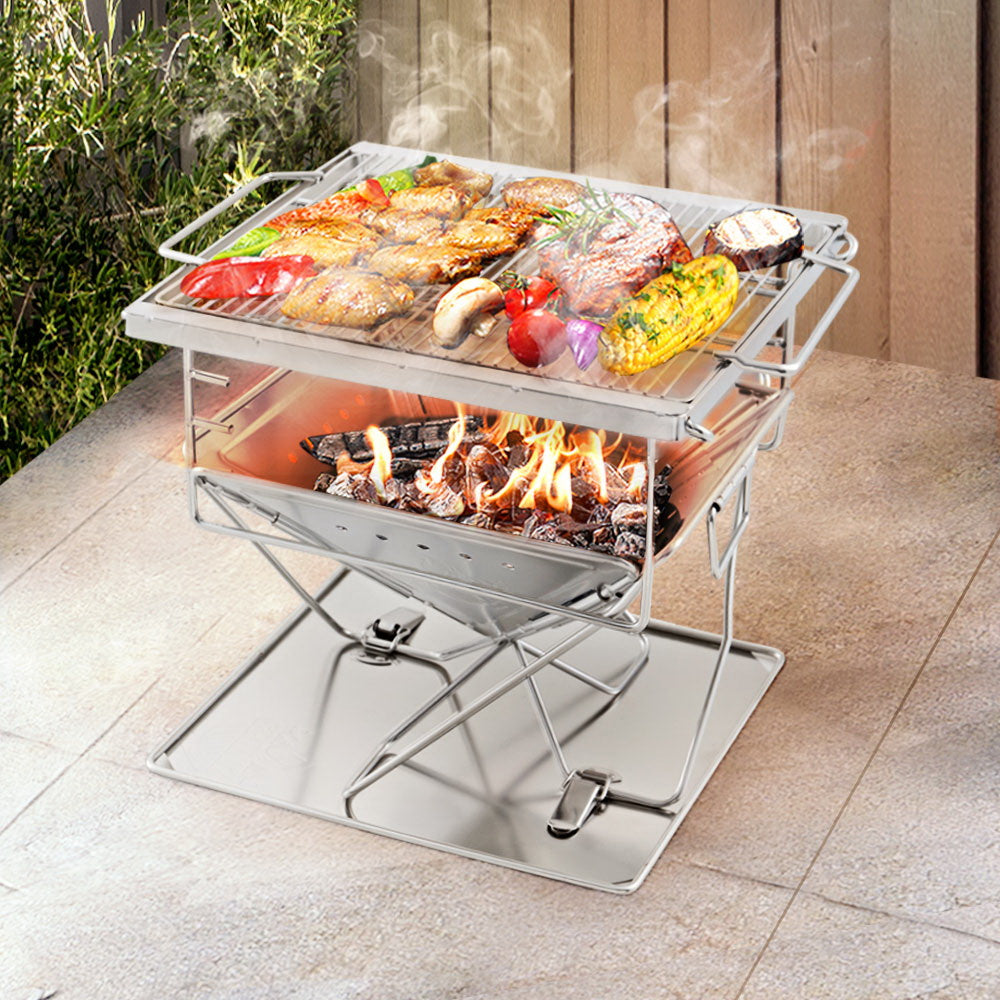 A portable stainless steel Grillz 2-in-1 Fire Pit BBQ Grill, cooking steak, chicken wings and vegetables set on an outdoor tiled surface in front of a wooden fence with garden setting with plants to the side.
