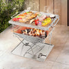 A portable stainless steel Grillz 2-in-1 Fire Pit BBQ Grill, cooking steak, chicken wings and vegetables set on an outdoor tiled surface in front of a wooden fence with garden setting with plants to the side.
