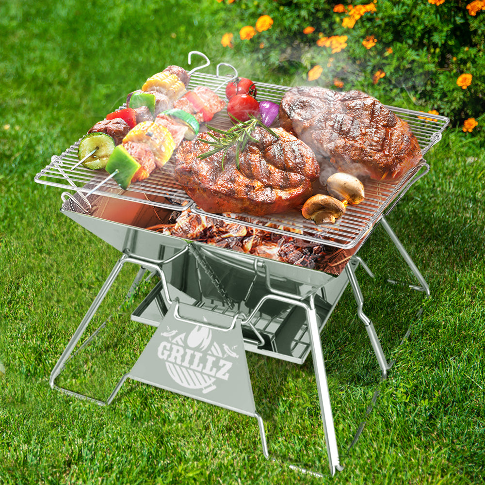 A portable stainless steel fire pit BBQ grill with a couple of steaks, skewered kebabs, tomatoes and mushrooms grilling on its grill rack. On a green grass area with marigolds in the background.