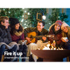 A group of five young rugged up people sitting around a traditional round rustic Fire Pit with a fire burning. There is a man playing a guitar, 3 women with wool beanies, holding mugs and another man with his arms crossed. All are happily smiling with a house, trees and hanging lights in the background.

