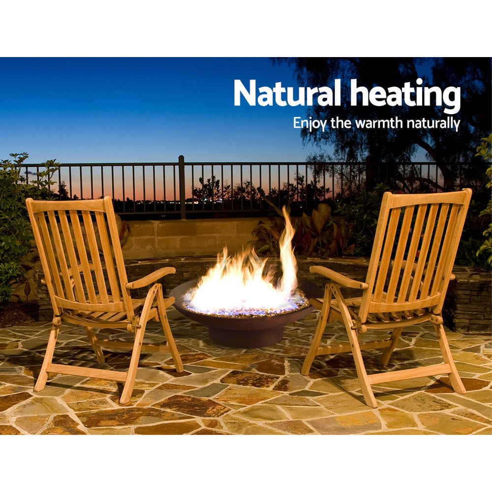 Grillz traditional round Fire Pit Bowl in Rust with fire burning and a couple of wooden deck chairs set on an outdoor slate floor area in front of a brick wall and fence with plants surrounding and a sunsetting view in the background.