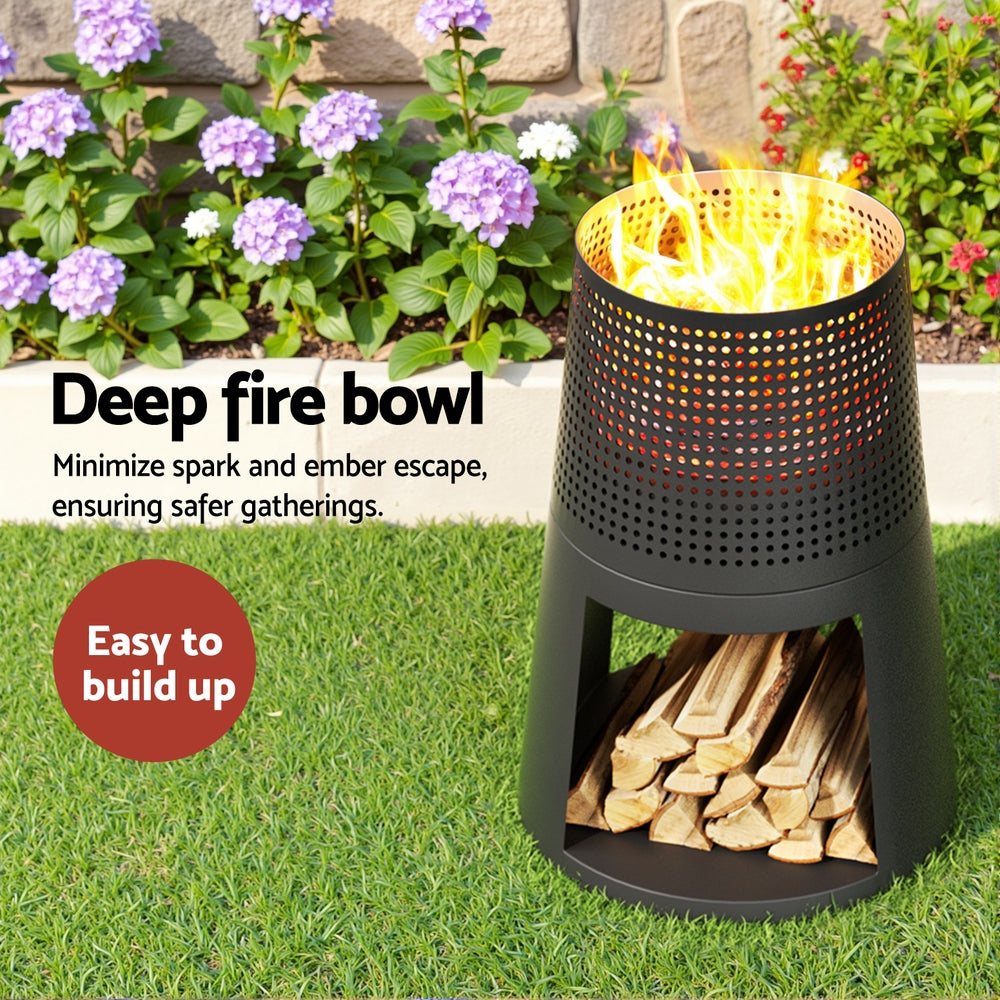 Black deep fire bowl with flames and firewood on grass, surrounded by flowers and a stone wall.