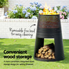 Black outdoor fire pit with wood storage on grass, surrounded by flowers.