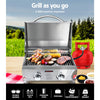 A portable stainless steel Grillz gas BBQ grill set on an outdoor table with steaks, kebabs, and vegetables cooking with a gas cylinder, tea towel and condiments on the table; grass and trees in the background.