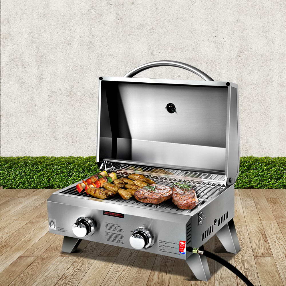 A portable stainless steel Grillz gas BBQ grill displayed in its opened configuration with steaks, chicken wings and kebabs cooking; set on a wood decking with a hedge and white wall in the background.