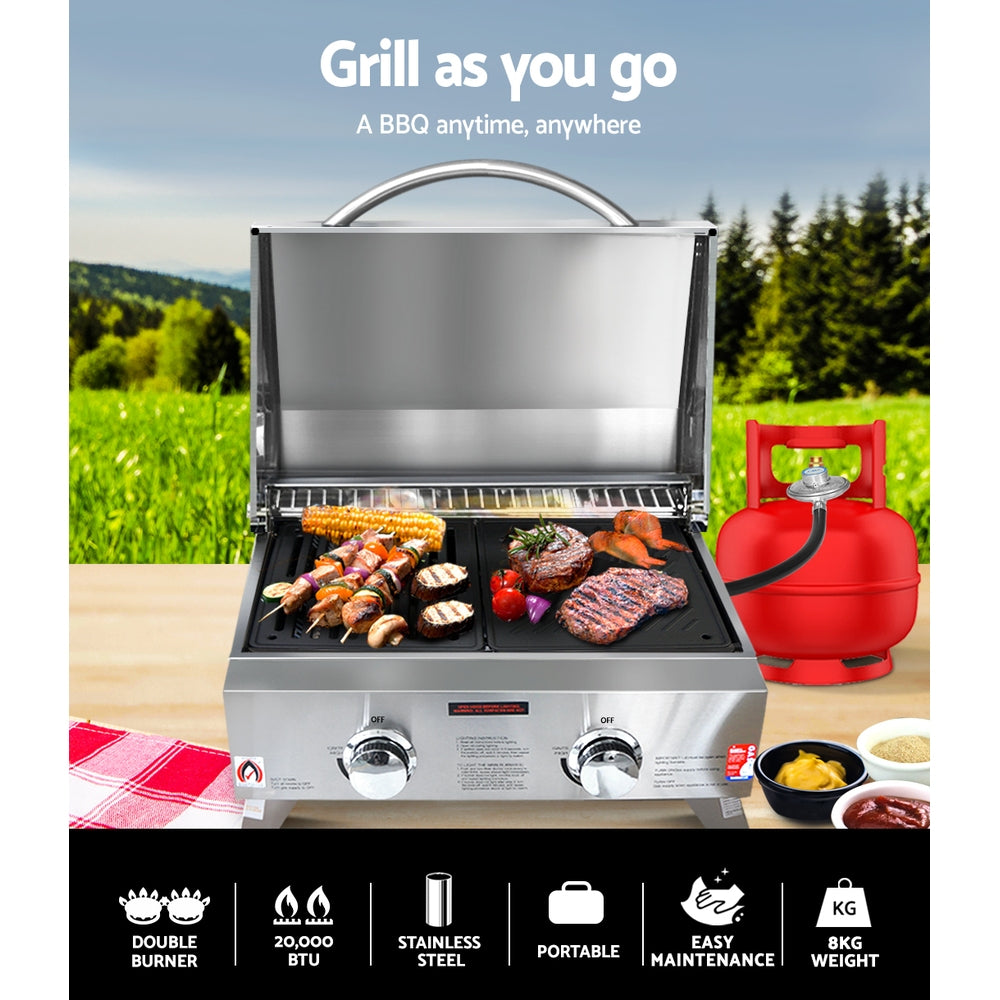 A portable stainless steel Grillz gas BBQ grill set on an outdoor table with steaks, kebabs, and vegetables cooking with a gas cylinder, tea towel and condiments on the table; grass and trees in the background.