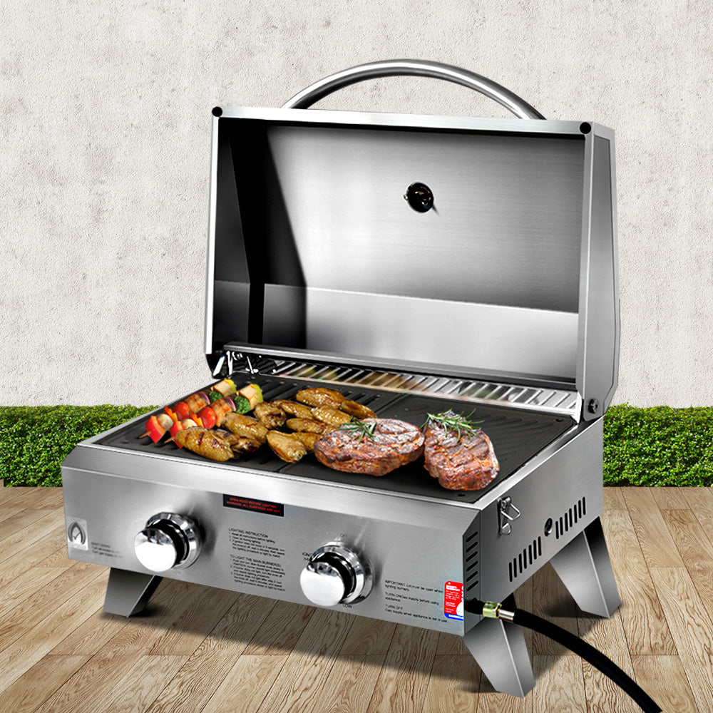 A portable stainless steel Grillz gas BBQ grill displayed in its opened configuration with steaks, chicken wings and kebabs cooking; set on a wood decking with a hedge and white wall in the background.
