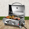 A portable stainless steel Grillz gas BBQ grill displayed in its opened configuration with steaks, chicken wings and kebabs cooking; set on a wood decking with a hedge and white wall in the background.
