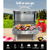 A portable stainless steel Grillz gas BBQ grill set on an outdoor table with steaks, kebabs, and vegetables cooking with a gas cylinder, tea towel and condiments on the table; grass and trees in the background.