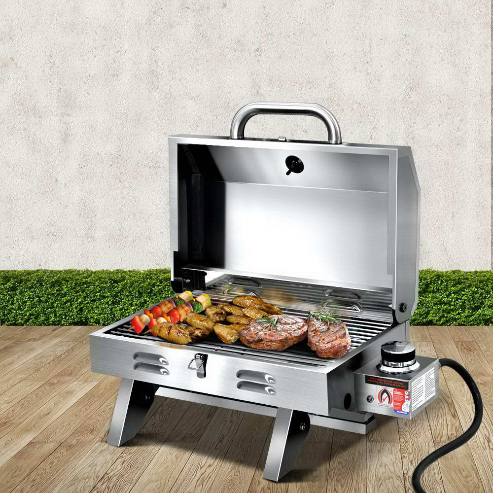 A portable stainless steel Grillz gas BBQ grill displayed in its opened configuration with steaks, chicken wings and kebabs cooking; set on a wood decking with a hedge and white wall in the background.