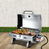 A portable stainless steel Grillz gas BBQ grill displayed in its opened configuration with steaks, chicken wings and kebabs cooking; set on a wood decking with a hedge and white wall in the background.