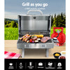 A portable stainless steel Grillz gas BBQ grill set on an outdoor table with steaks, kebabs, and vegetables cooking with a gas cylinder, tea towel and condiments on the table; grass and trees in the background.