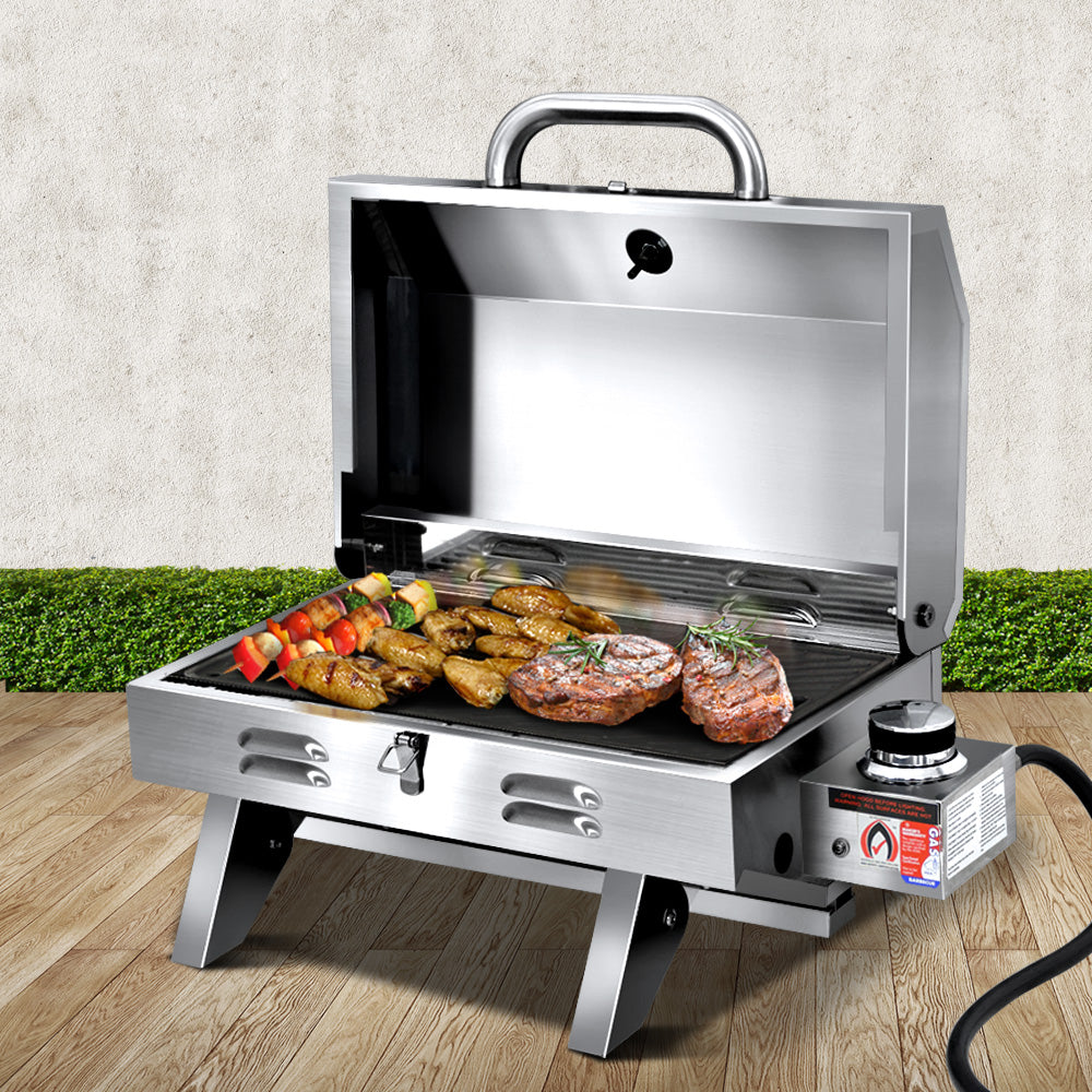 A portable stainless steel Grillz gas BBQ grill displayed in its opened configuration with steaks, chicken wings and kebabs cooking; set on a wood decking with a hedge and white wall in the background.