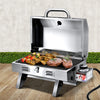A portable stainless steel Grillz gas BBQ grill displayed in its opened configuration with steaks, chicken wings and kebabs cooking; set on a wood decking with a hedge and white wall in the background.