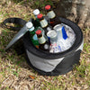 Portable cooler with drinks and ice set up on grass area beside a tree stump.
