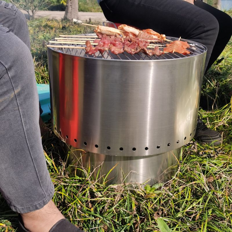 A stainless-steel Inferno smokeless fire pit, shown ready to cook kebabs and chicken in an outdoor setting.