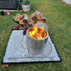 A stainless steel Inferno smokeless fire pit with fire burning with a grill, poker and log stack on a gravel setting in a private backyard.