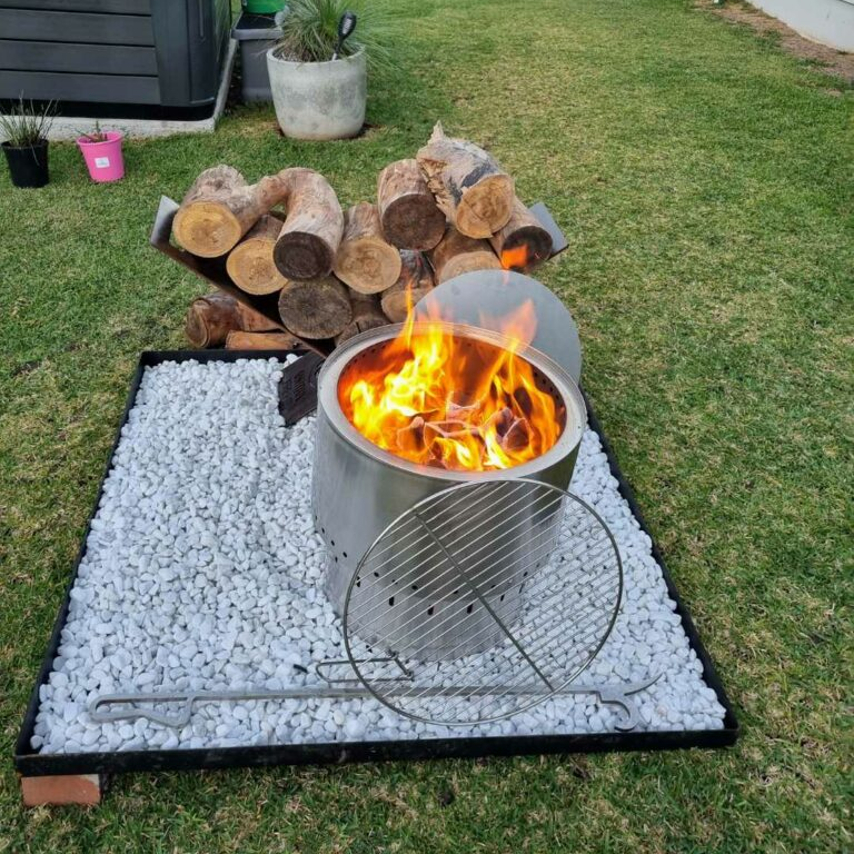A stainless steel Inferno smokeless fire pit with fire burning with a grill, poker and log stack on a gravel setting in a private backyard.