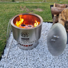 A stainless steel Inferno smokeless fire pit with fire burning with a grill leaning on it, a poker, lid and logs stacked on a gravel setting in a backyard.