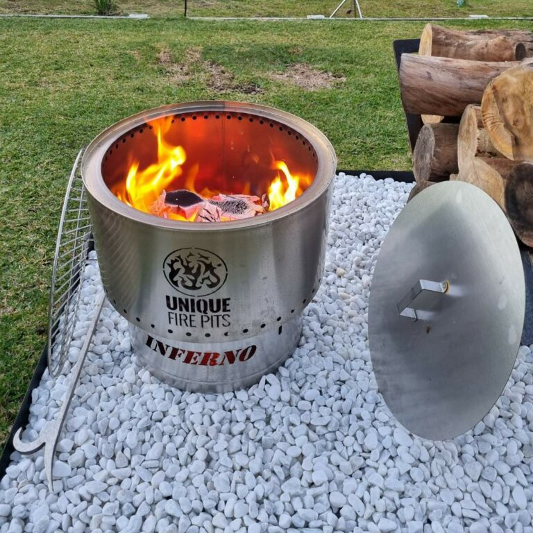 A stainless steel Inferno smokeless fire pit with fire burning with a grill leaning on it, a poker, lid and logs stacked on a gravel setting in a backyard.