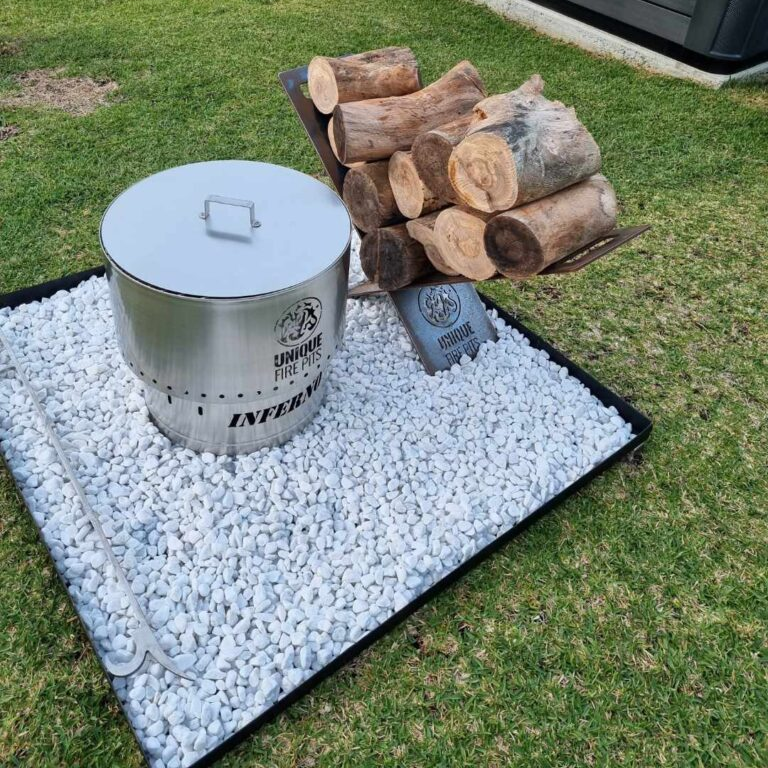 A stainless steel Inferno smokeless fire pit with lid on it, a poker beside it, and logs stacked on a log rack on a gravel setting in a backyard.