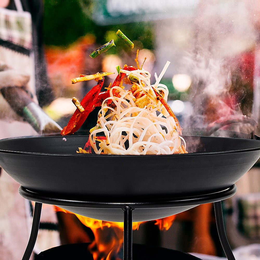 A matte black iron fire bowl with a traditional and minimalist design, supported by three curved legs being used as a wok, with noodles and vegetables being stir fried above a burning flame.