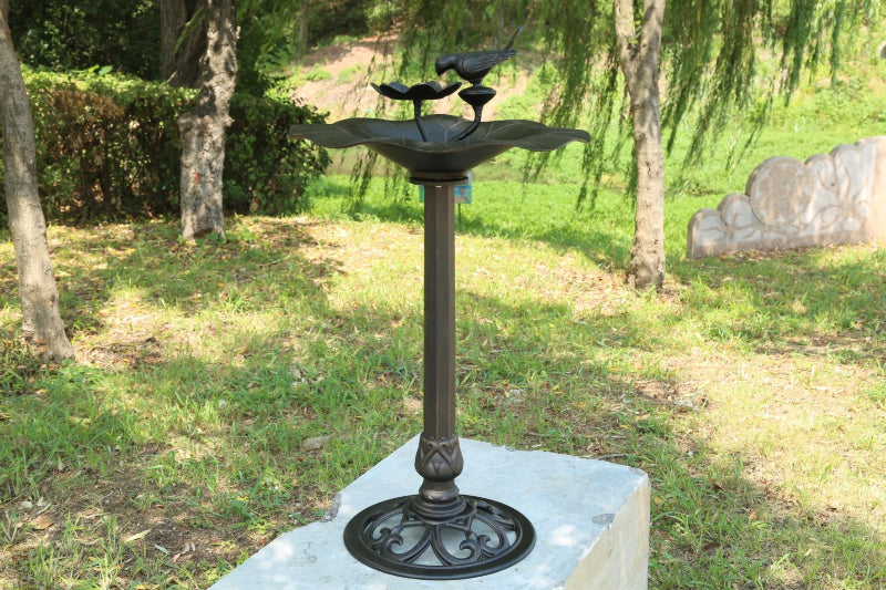 Decorative outdoor bird bath in a garden setting on a cement block with grass, trees, hedge and a cement wall in the backdrop.