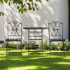 Black metal garden table and chairs on a grassy lawn with a white wall and greenery in the background.