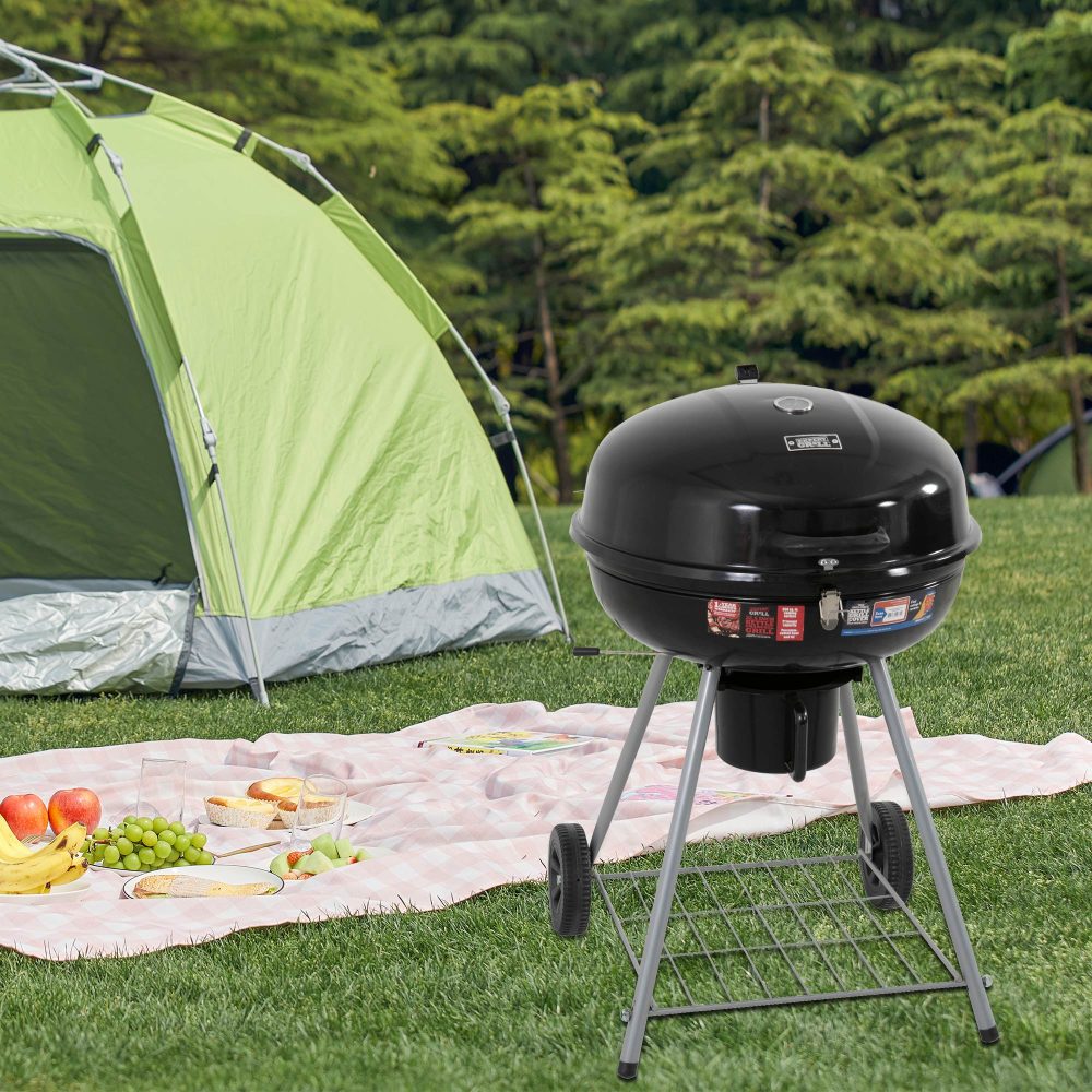 A portable charcoal BBQ smoker with a black enameled firebowl, lid, chrome plated vent discs, and a tripod design. set in front of a tent and picnic on grassed area with trees in the backdrop.