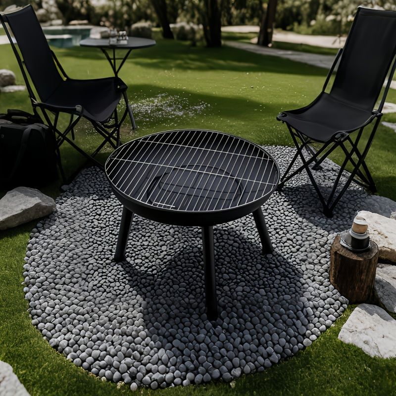 Black outdoor grill on a pebble circle with chairs and a table in a garden setting