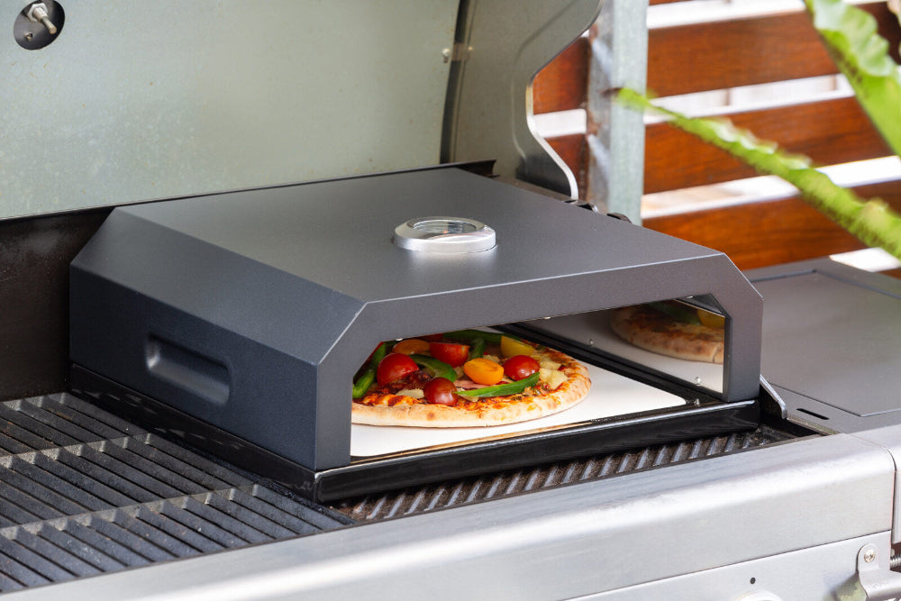 Portable pizza oven on a grill with a pizza inside, surrounded by outdoor elements.