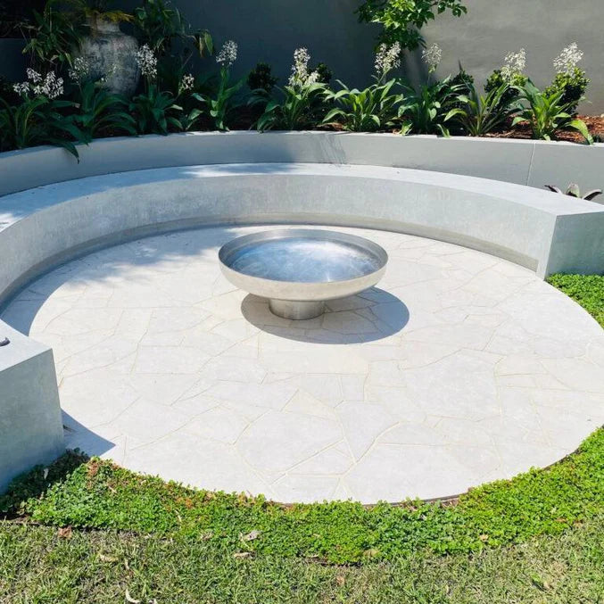 Cauldron round stainless-steel fire pit on cement tiled area with cement circular seating within a grassed area and plants in the background.