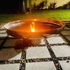 A polished mirror finished stainless steel fire pit with a circular shape, situated on a cement tiled area with a small fire burning and a lawn and plant background