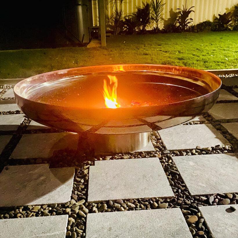 A polished mirror finished stainless steel fire pit with a circular shape, situated on a cement tiled area with a small fire burning and a lawn and plant background