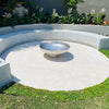A polished mirror finished stainless steel fire pit situated on a cemented area with cement bench seating and flowering plants in the background