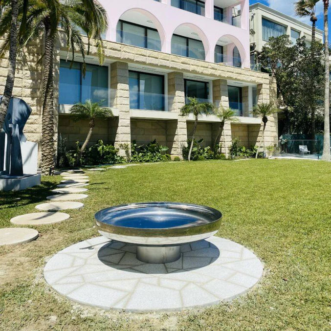 Cauldron round stainless-steel fire pit on cement tiled area with a lawned area and building in the background.