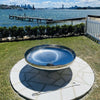 Cauldron round stainless-steel fire pit on a cement tiled area with a grass and hedging at a seaside with jetty, boats and buildings in the background.