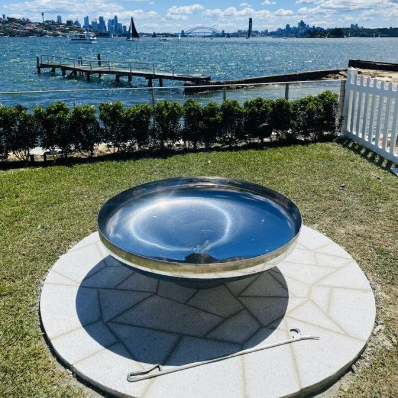 Cauldron round stainless-steel fire pit on a cement tiled area with a grass and hedging at a seaside with jetty, boats and buildings in the background.