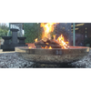 A round polished mirror finish stainless steel fire pit with a burning log fire, on a gravelled area with house, water feature and plants in the background.