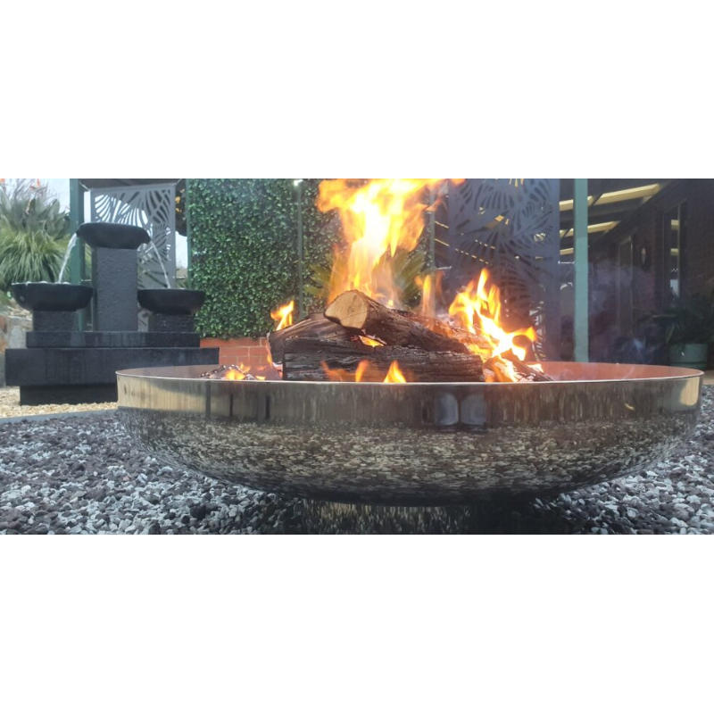 A round polished mirror finish stainless steel fire pit with a burning log fire, on a gravelled area with house, water feature and plants in the background.