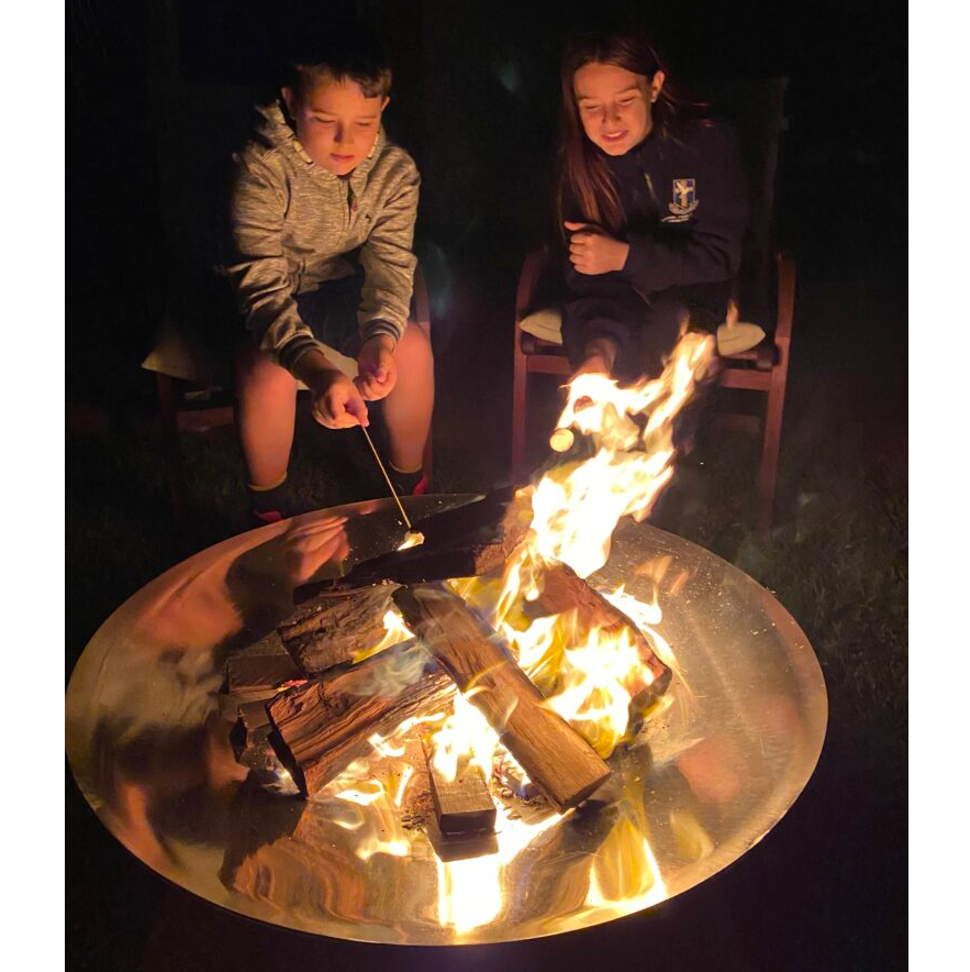 Two children sitting beside a round shaped polished mirror finished stainless steel fire pit and toasting marshmellows.