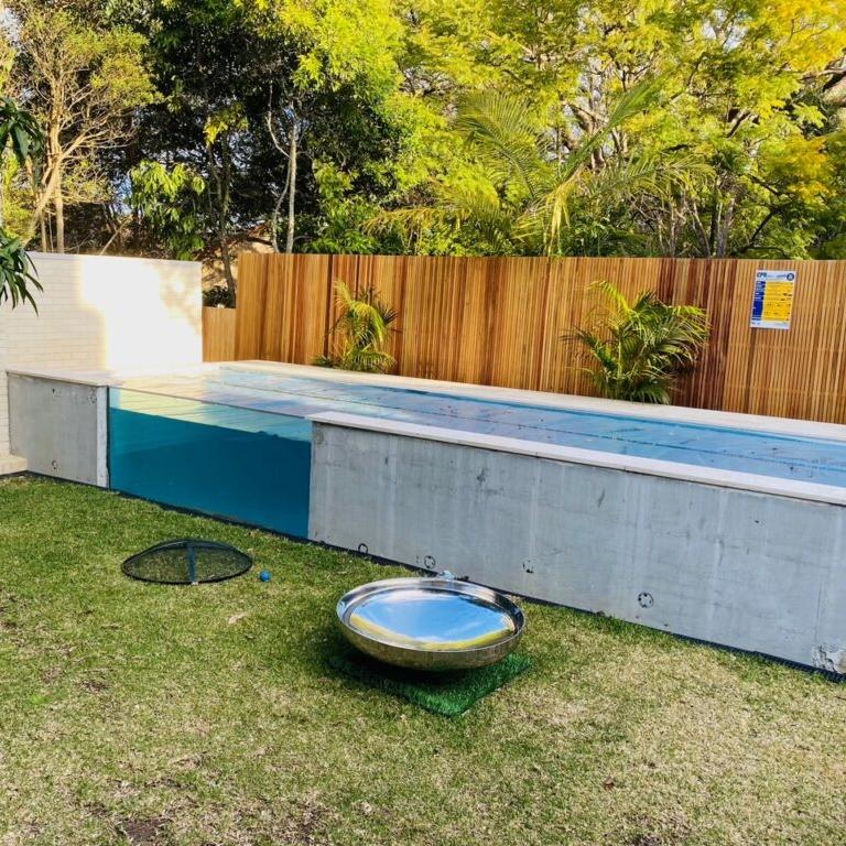 A polished mirror finished stainless steel fire pit situated on a lawn area with an ember screen next to it and beside a lap pool with trees in the background