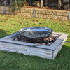 A polished mirror finish stainless steel fire pit with a circular shape, situated in a garden setting with grass and plants in the background.