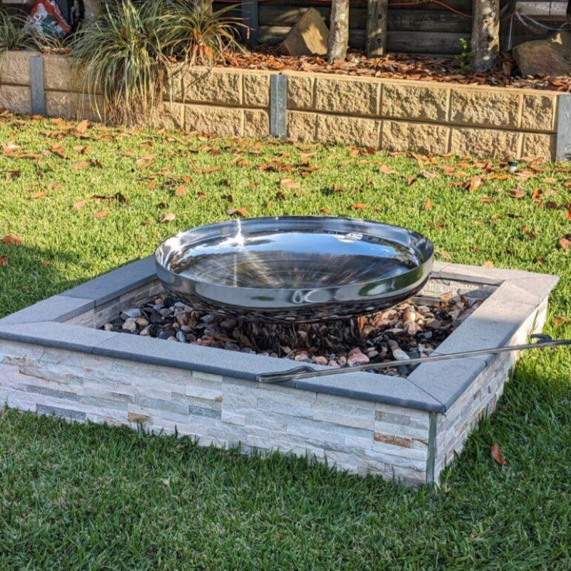A polished mirror finish stainless steel fire pit with a circular shape, situated in a garden setting with grass and plants in the background.