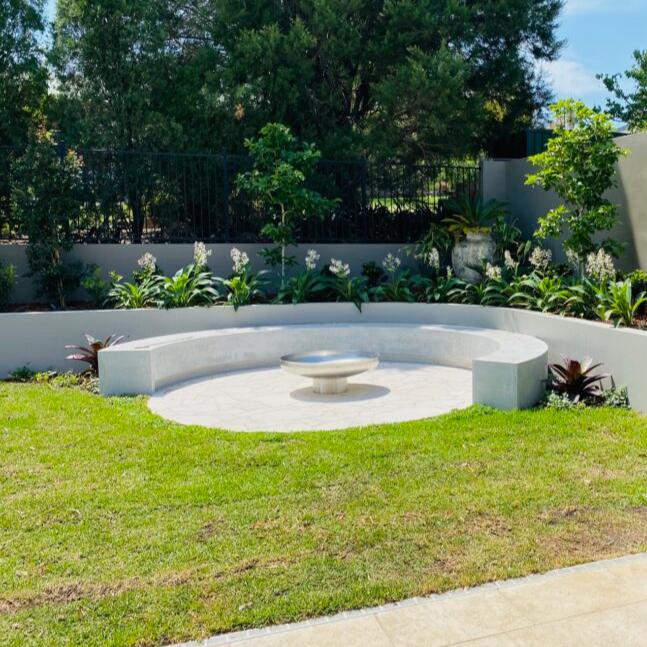 A polished mirror finished stainless steel fire pit situated on a cemented area in lawned garden setting with cement bench seating and flowering plants and trees in the background 